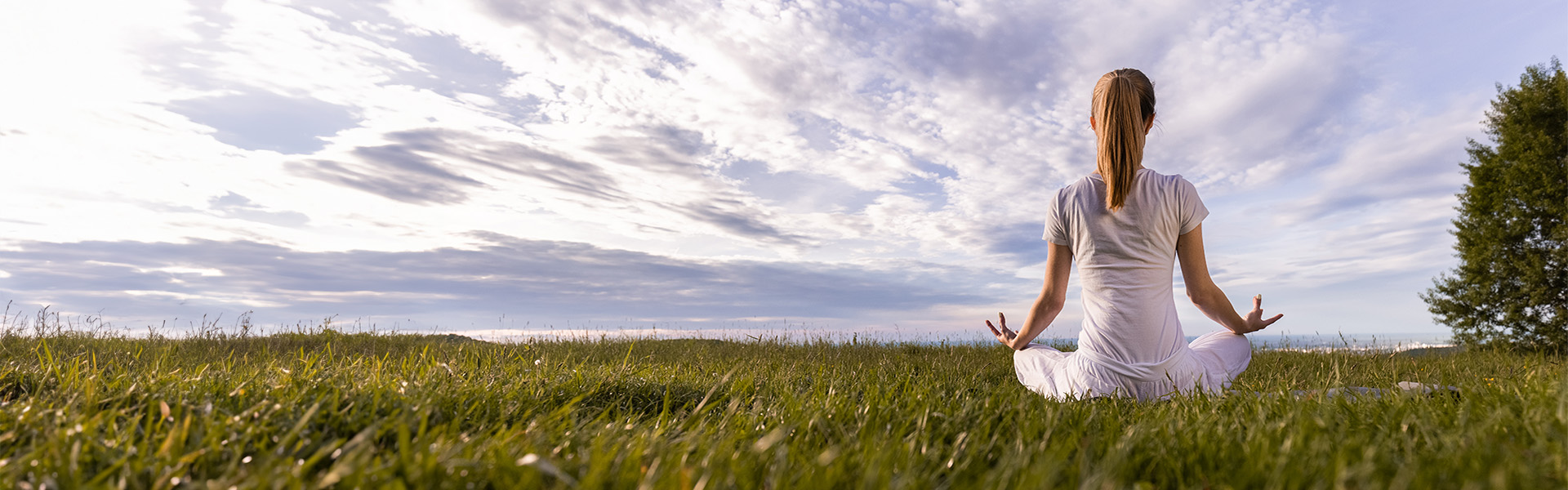 Woman sitting on the grass in a meditation pose with blue sky and clouds in the distance.
